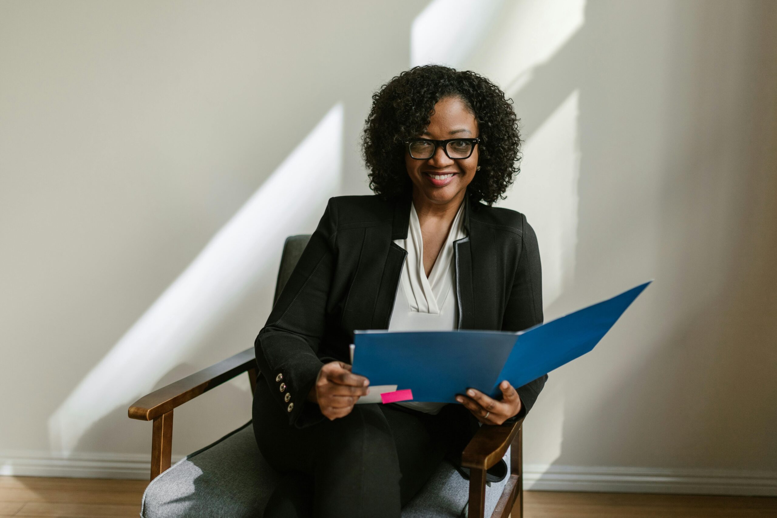 Professional African American woman with glasses and curly hair holding a blue folder indoors.